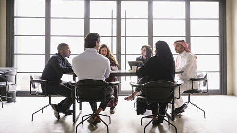 A group of people sitting around a table in a meeting room with large windows in the background. Some are using laptops and taking notes.