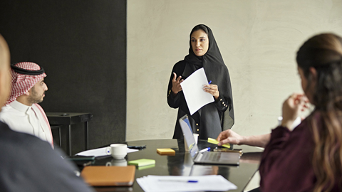 A person in a black outfit is standing and holding papers, speaking to a group of people seated around a table with laptops, notebooks, and coffee cups.